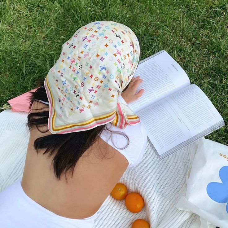Person in floral bucket hat reading book on grass with white blanket and sunglasses