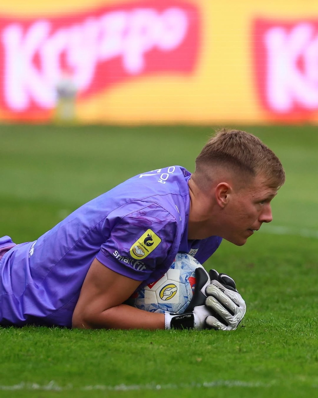 Soccer goalkeeper in a purple uniform lying on grass while holding a ball
