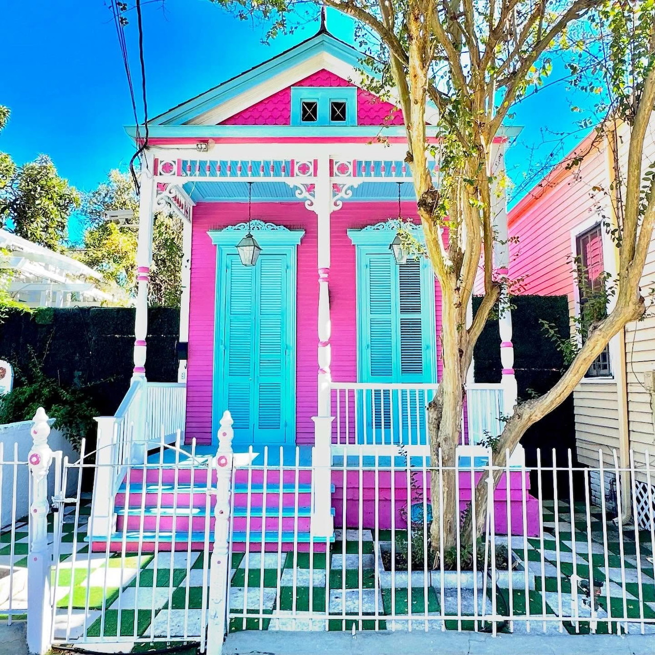 Bright pink cottage with turquoise door, white trim, and colorful striped porch behind white picket fence