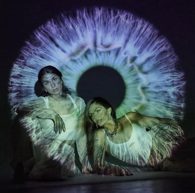 Two women posing in front of a large glowing eye projection in purple and teal tones on a dark background