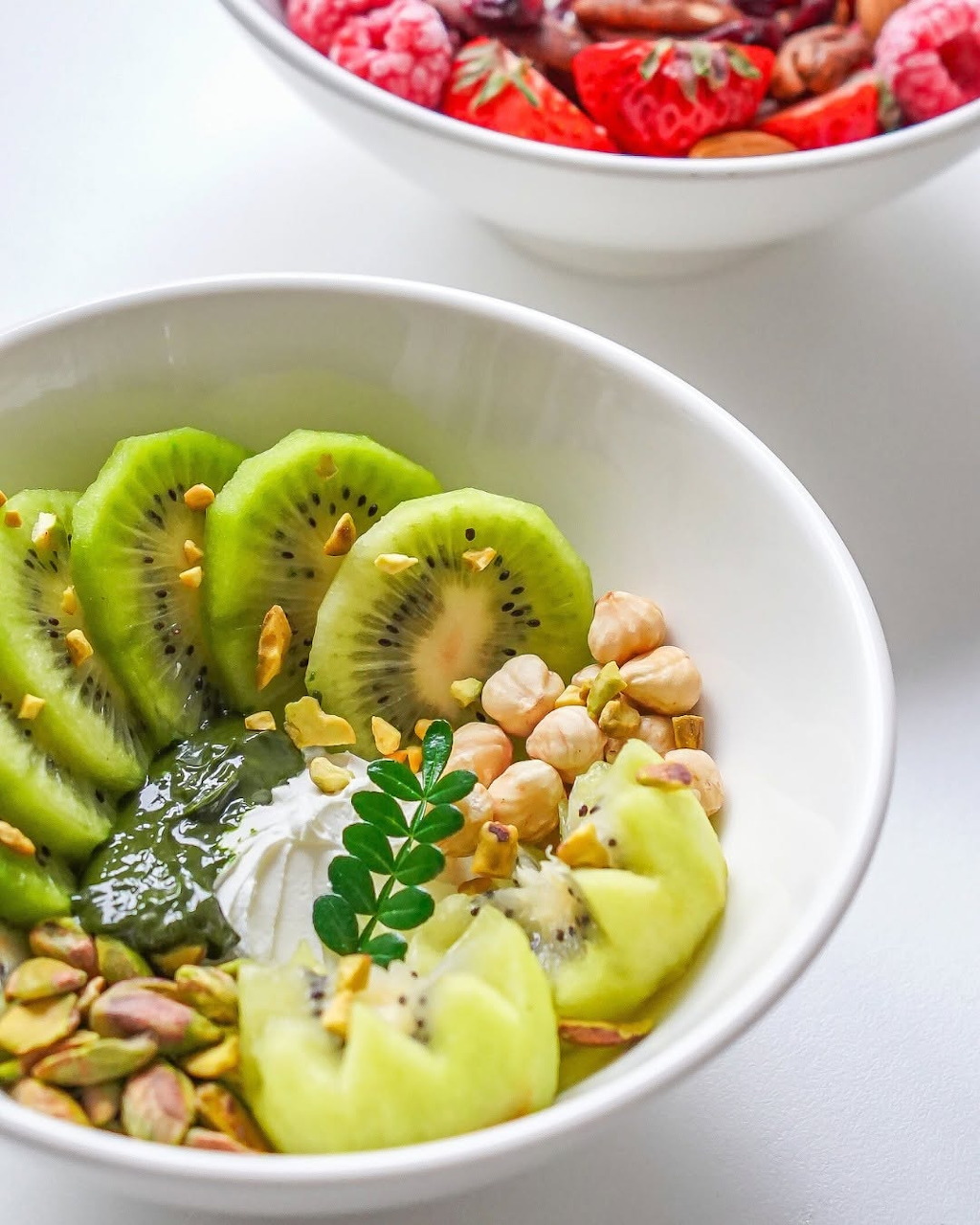 White bowl with sliced kiwi, yogurt, pistachios, and seeds on a white surface
