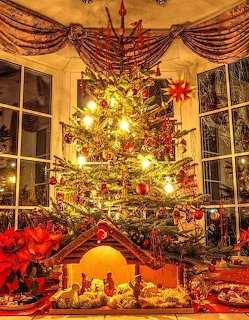 Illuminated Christmas tree with red ornaments, nativity scene below, and poinsettias in a warm indoor setting