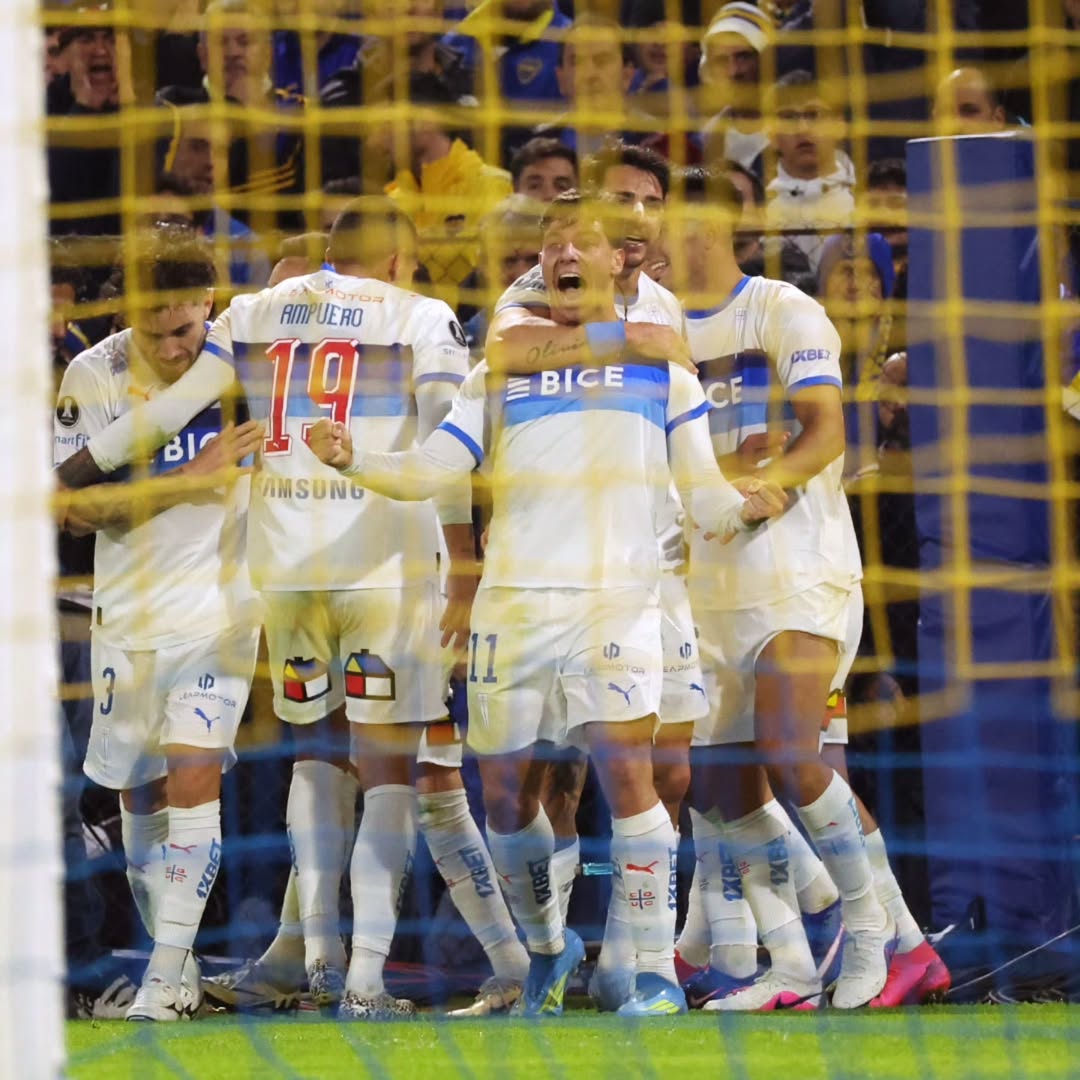 Soccer players in white kits celebrating together behind a yellow goal net