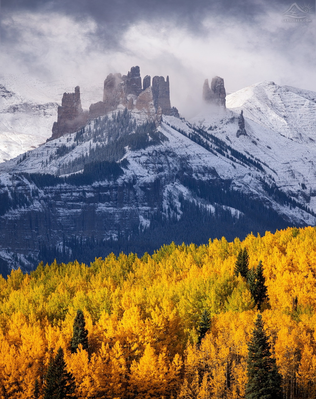 Rocky mountain peaks shrouded in clouds rising above golden aspen trees in autumn