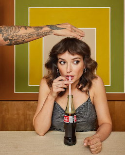 Woman with curly brown hair drinking from a glass bottle with a straw at a table