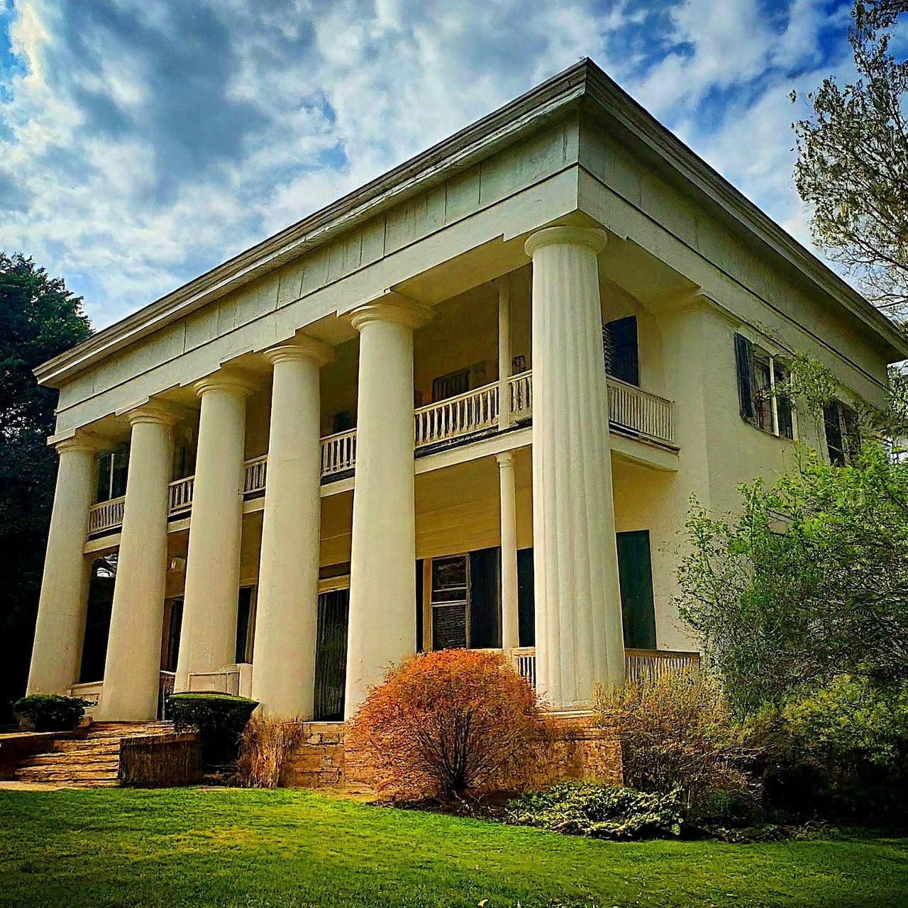 Greek Revival mansion with tall white columns and yellow facade on green lawn under cloudy blue sky