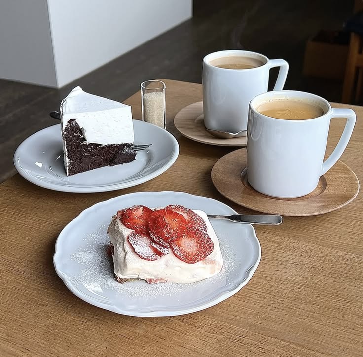 Two coffee mugs with strawberry cake and chocolate cake on plates at a cafe table