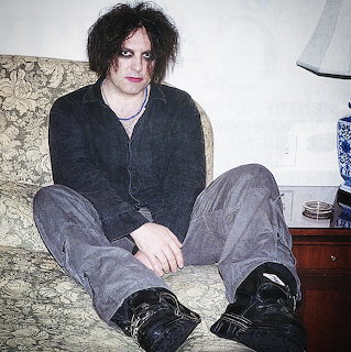 Robert Smith seated on a patterned sofa indoors, wearing dark clothes and black shoes