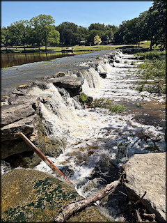 Wide cascading waterfall over rocky ledges in a green park with trees on a sunny day