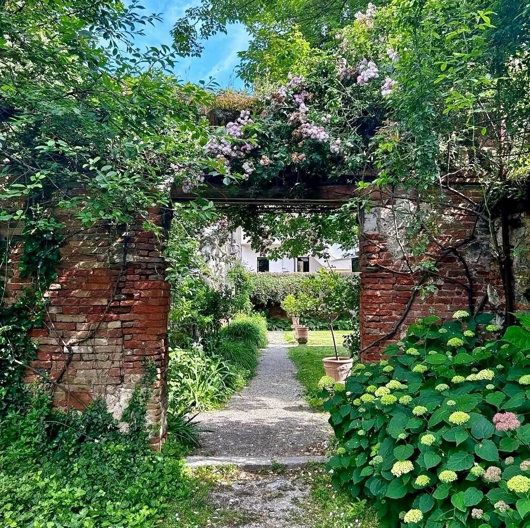 Stone archway with white flowers opening to garden path with hydrangeas and brick walls