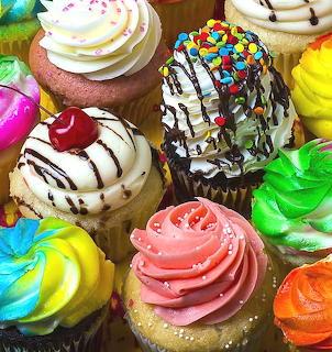 Assorted cupcakes with colorful swirled frosting, chocolate sauce, candy sprinkles, and cherry topping
