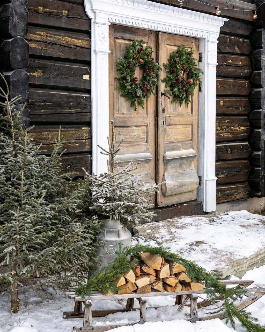 Wooden cabin door with Christmas wreaths, small evergreen tree, firewood stack, and snow on ground