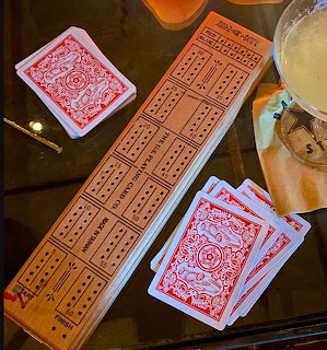 Wooden cribbage board with peg holes and two stacks of red playing cards on a glass table