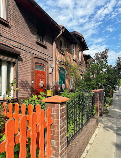 Brick row houses with red and teal doors behind an orange wooden picket fence and green plants