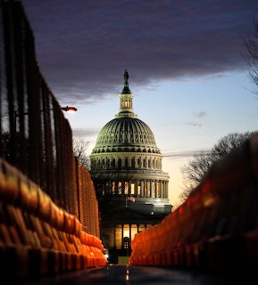 Illuminated US Capitol dome at dusk framed by orange barriers and dark fencing