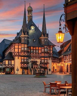 Historic half-timbered buildings in a cobblestone town square at dusk with a glowing street lantern