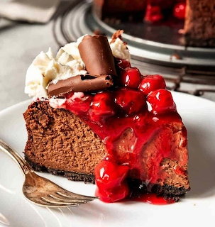 Slice of chocolate cheesecake with cherry topping, whipped cream, and chocolate curls on white plate