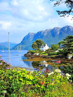 White cottage by a mountain lake with sailboat, rugged peaks, and colorful garden in foreground