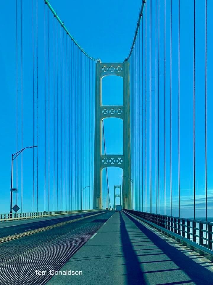 Road deck perspective of Mackinac Bridge with tall suspension towers against a bright blue sky
