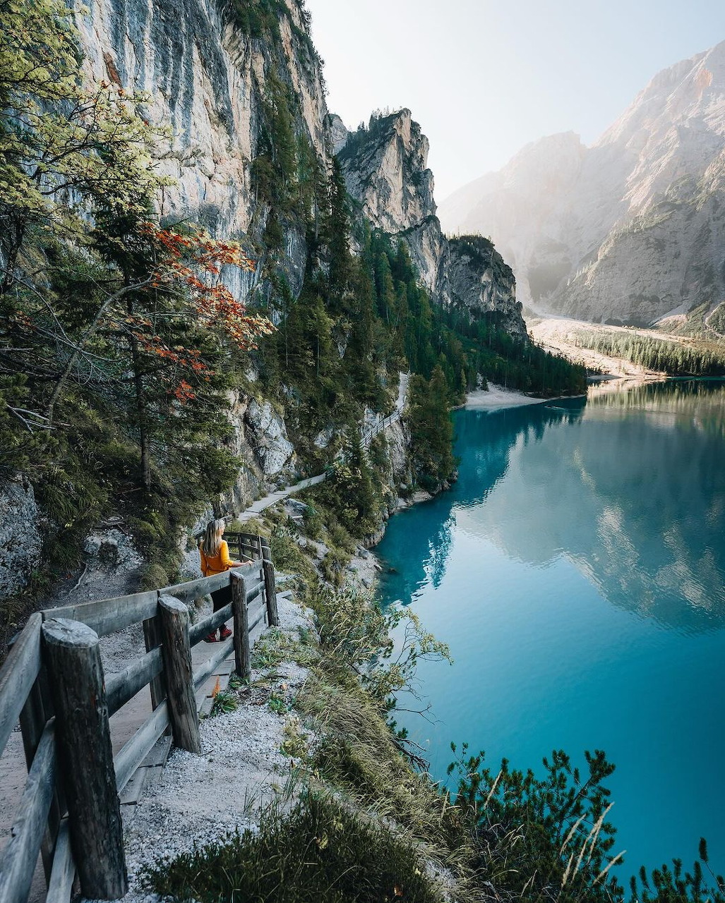 Hiker on wooden boardwalk trail along rocky cliffs overlooking a vivid turquoise alpine lake