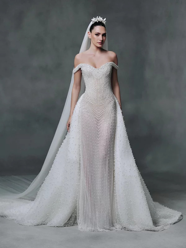 Model in off-shoulder beaded white wedding dress with long veil and tiara against grey background