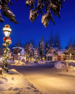 Snowy village street at dusk with glowing street lamps and festive lights under blue twilight sky