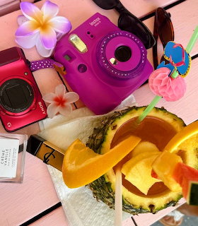 Flatlay of purple instant camera, yellow straw hat, pink flowers, and beach items on pink wood