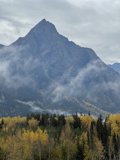 Tall mountain peak with mist and clouds above autumn forest with yellow trees and evergreens in British Columbia