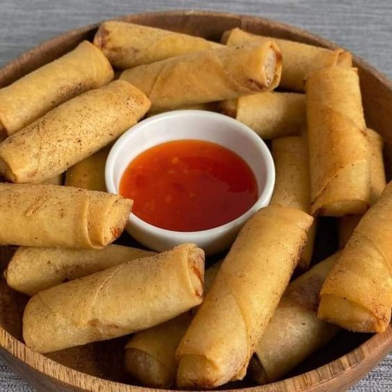 Crispy golden egg rolls in a wooden bowl with red sweet chili dipping sauce in a white dish