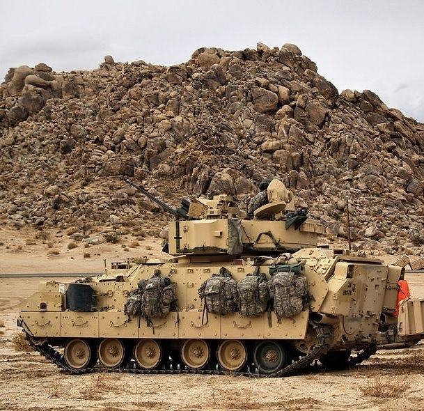 Tan Bradley armored vehicle parked on desert ground with a rocky hill behind it