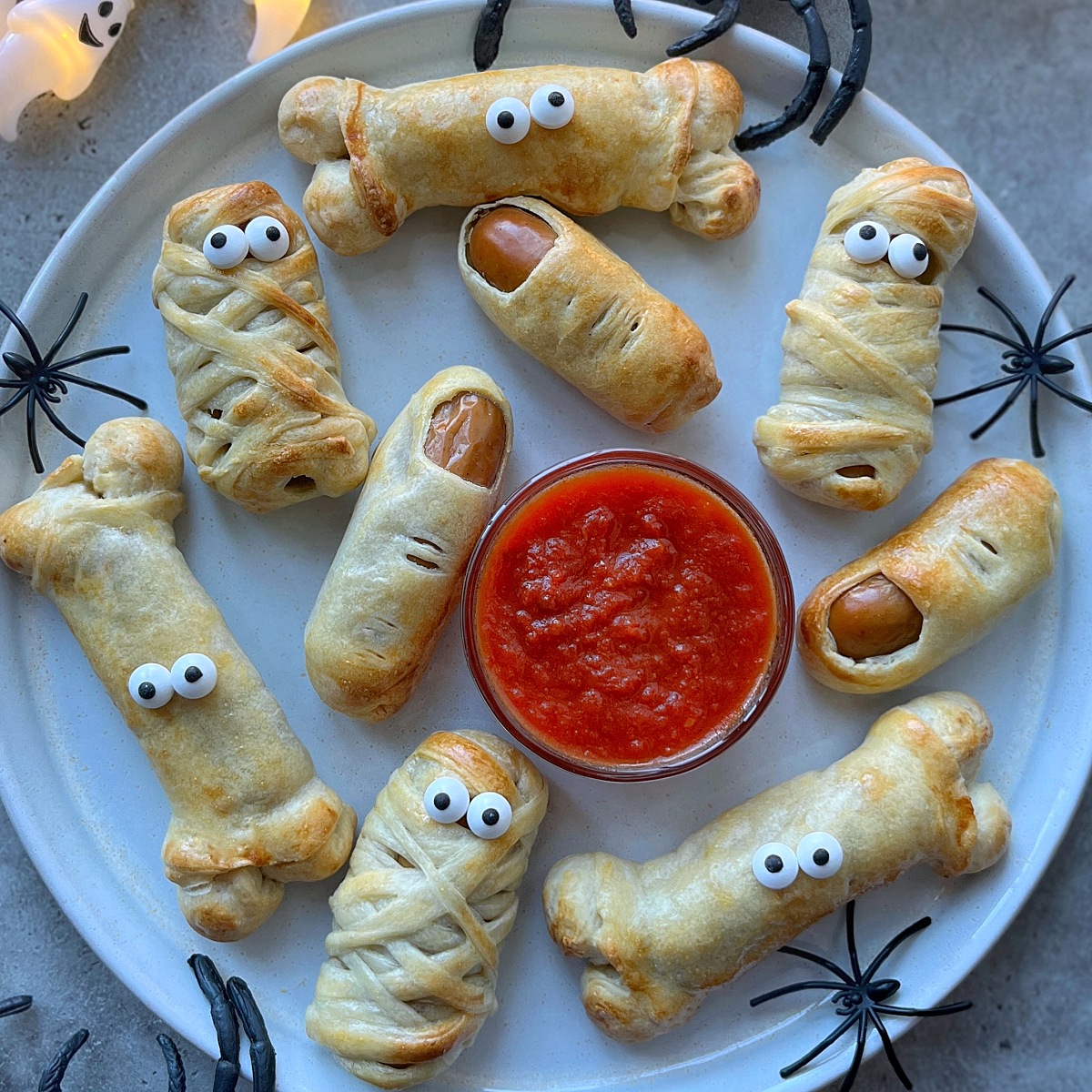 Plate of spooky baked finger and mummy snacks with googly eyes around red dipping sauce