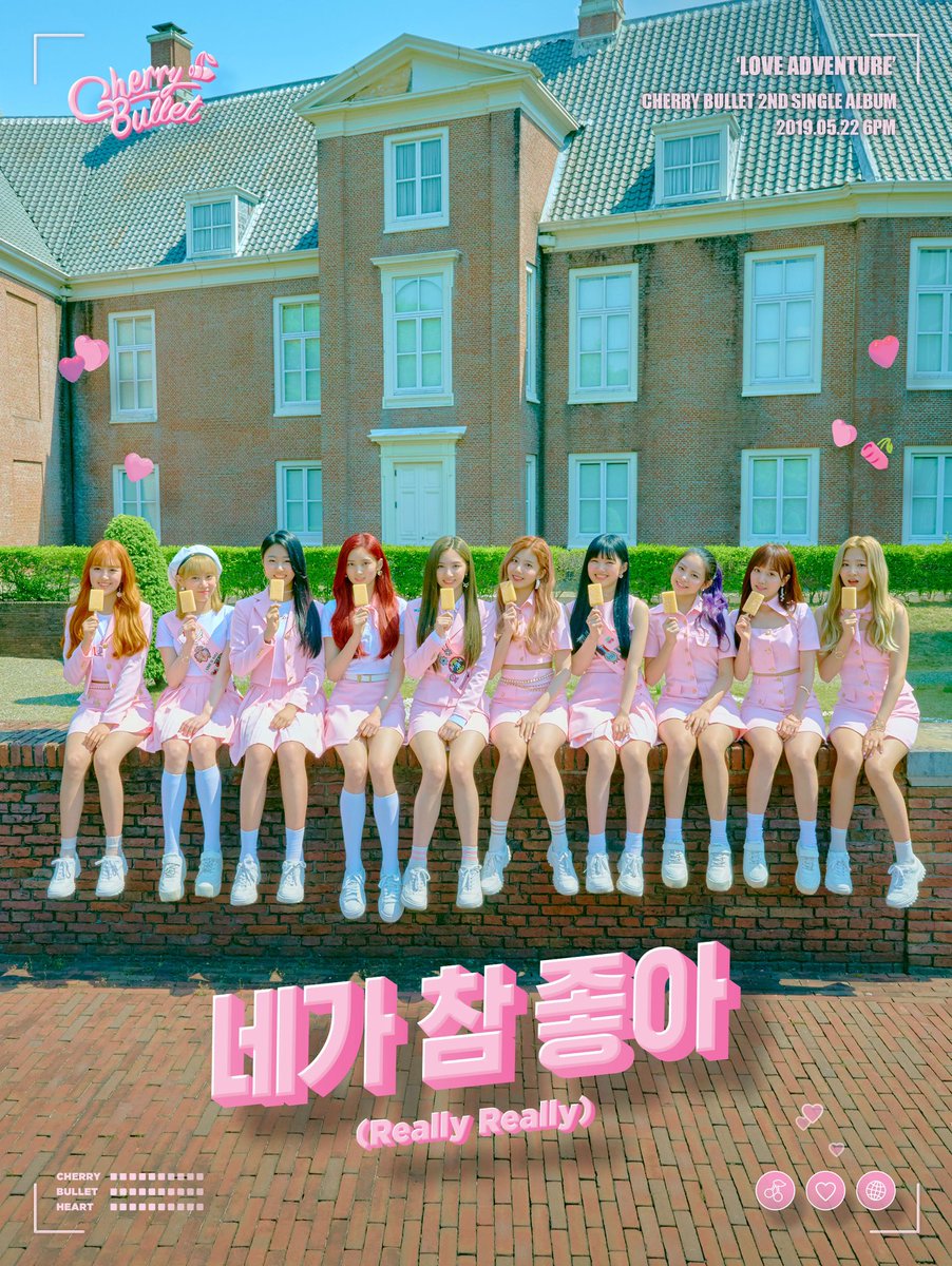 Cherry Bullet K-pop girl group in pink outfits sitting together outdoors in front of a brick building
