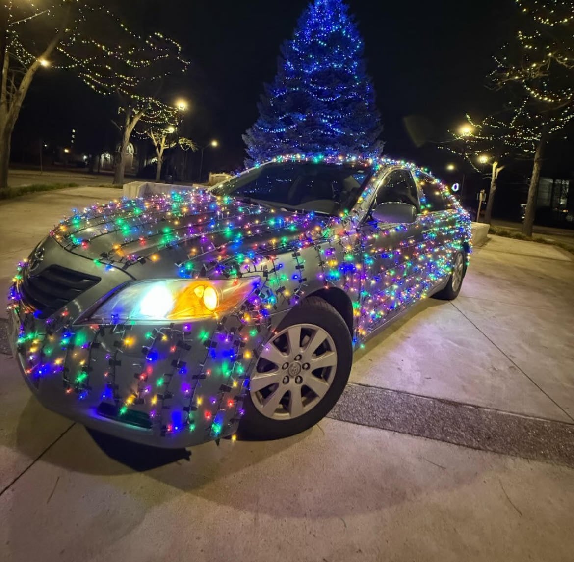 Silver car wrapped in colorful Christmas lights parked outdoors at night near lit trees