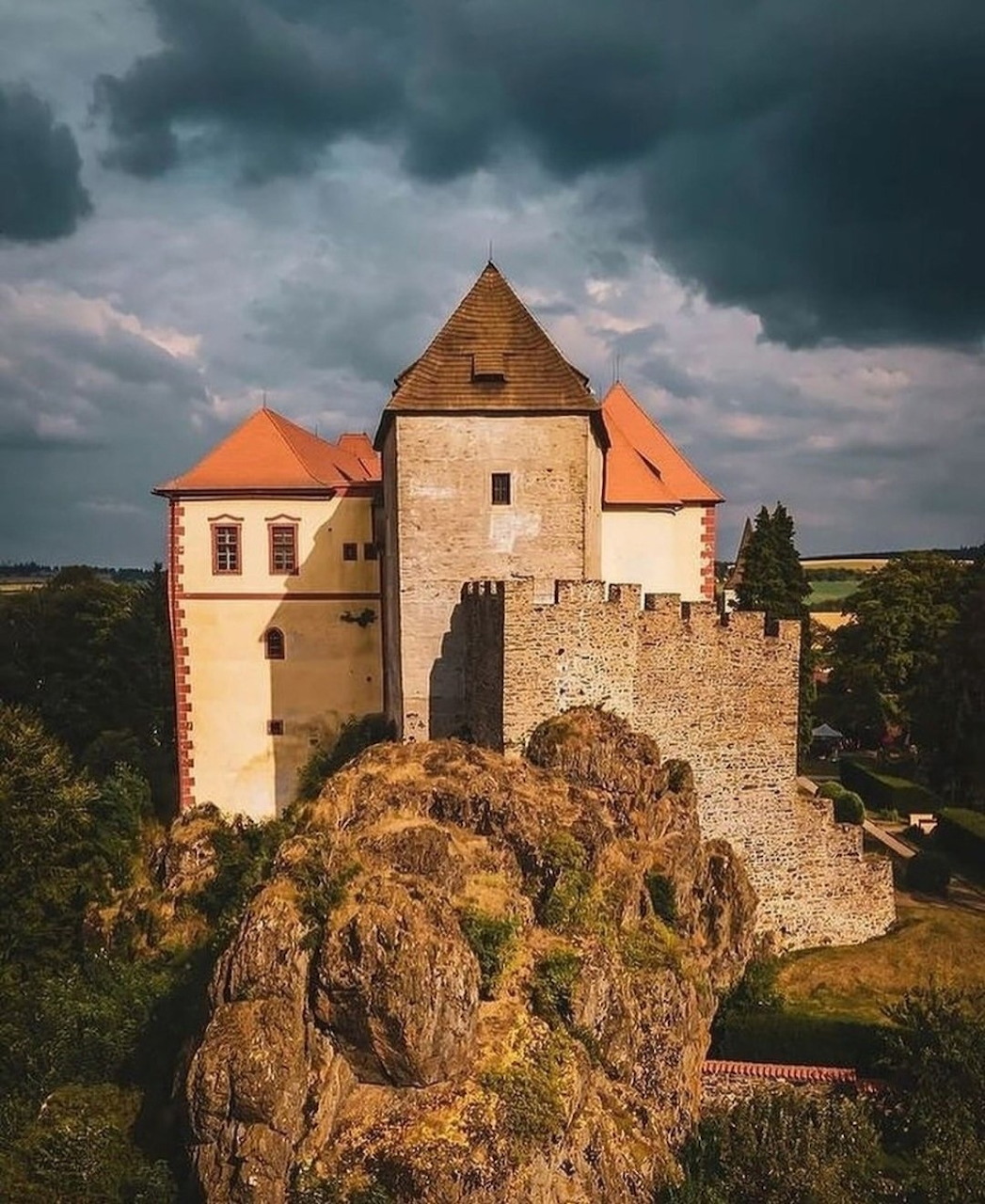 Medieval castle on rocky hilltop with orange roofs against stormy sky in Czech Republic