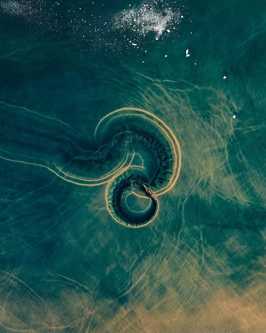 Aerial view of ocean wave forming spiral pattern with turquoise water and white foam against sandy bottom