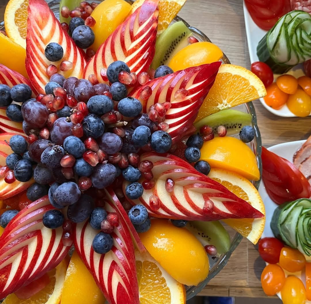 Ornate fruit platter with carved red apple petals, grapes, orange slices, kiwi, and berries arranged in a flower pattern