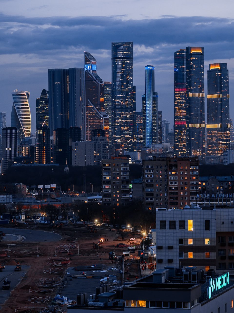 Moscow City skyscrapers lit up at dusk with dark clouds overhead and residential area in foreground