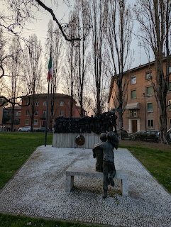 Bronze memorial sculpture with figures in a park plaza surrounded by bare trees and apartment buildings