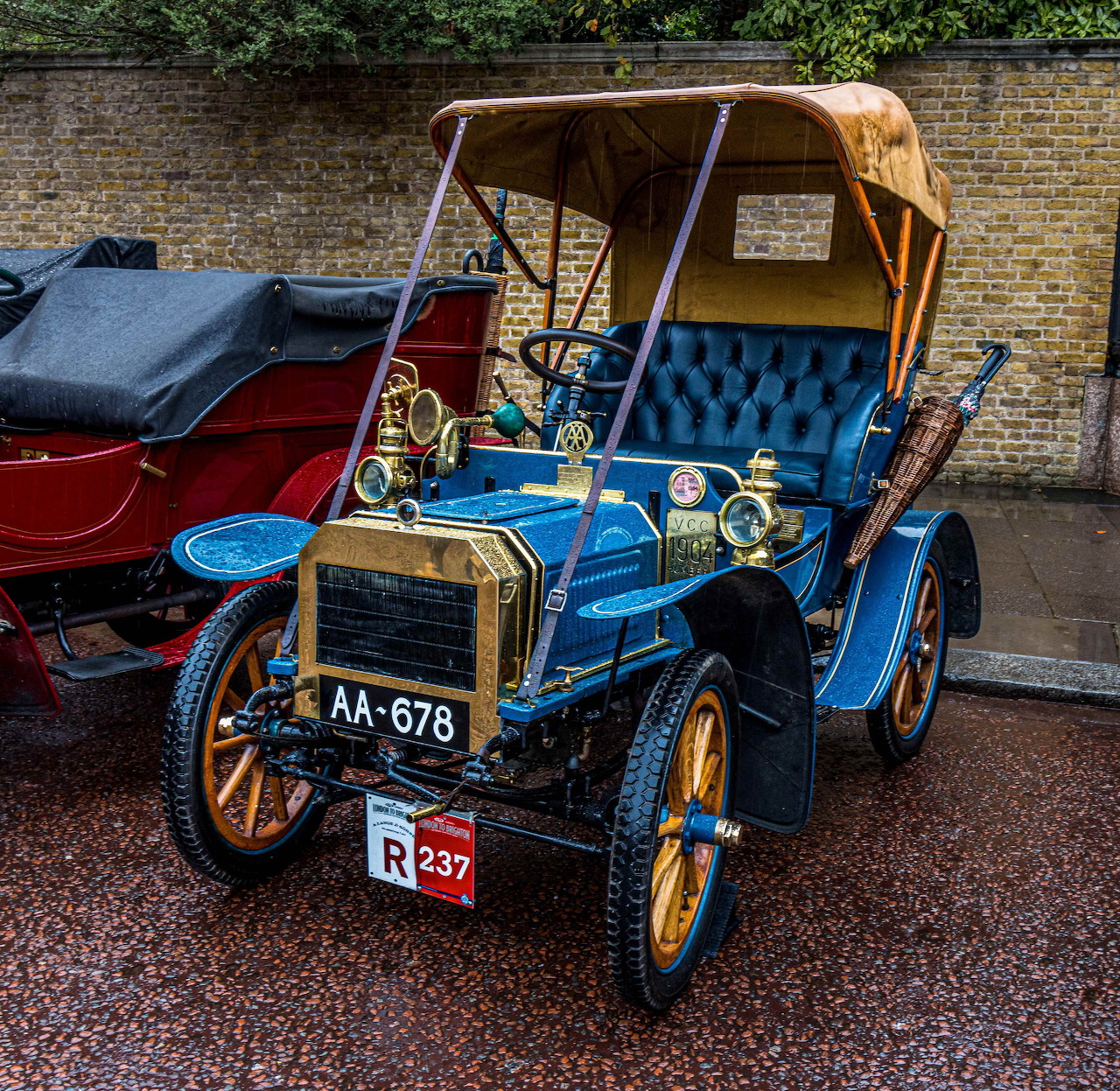 Vintage blue open-top car parked on a wet street beside a brick wall