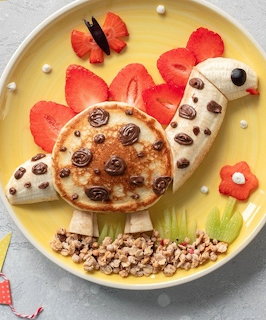 Stegosaurus made from pancake, banana slices, strawberries, chocolate chips, and cereal on a yellow plate