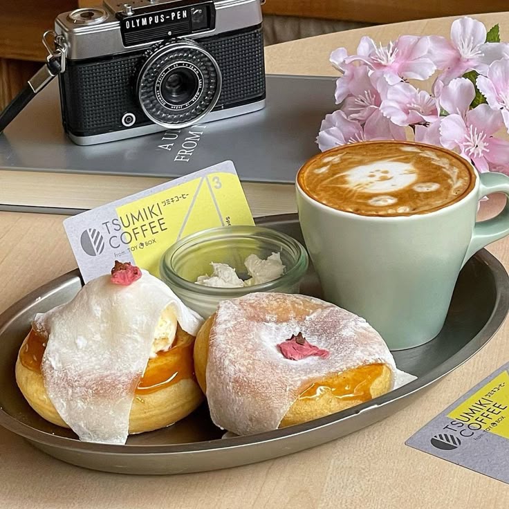 Latte and two pastries on a tray with a vintage camera and pink flowers in the background.