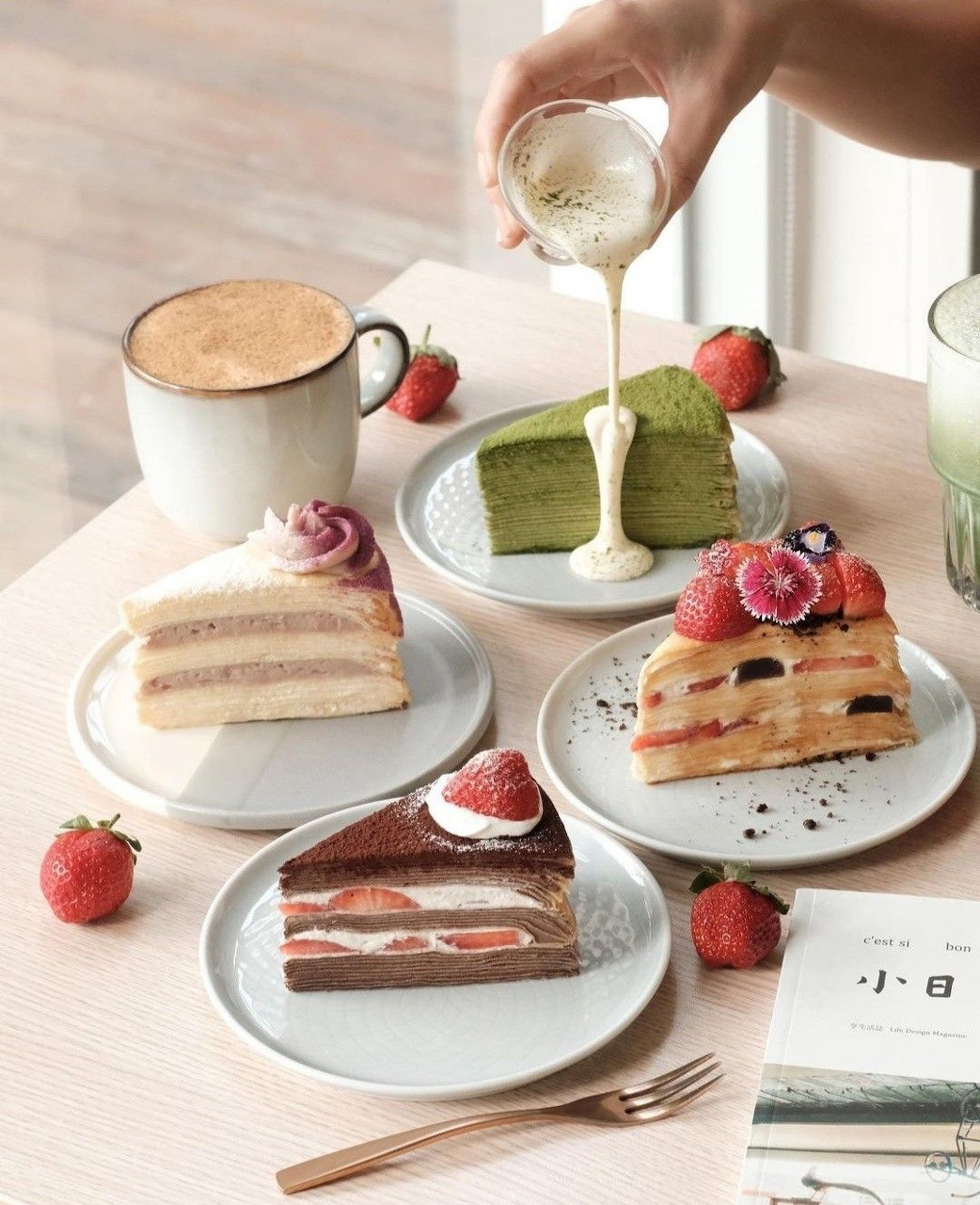Multiple layered cake slices on plates with coffee cups and strawberries, hand pouring cream