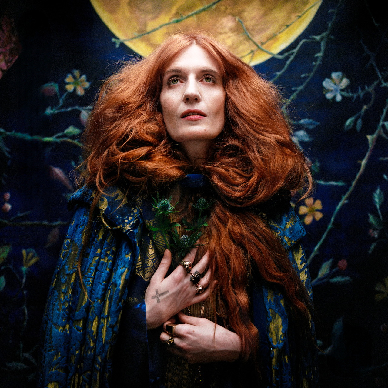 Woman with long red hair in blue embroidered garment against dark floral background with yellow moon