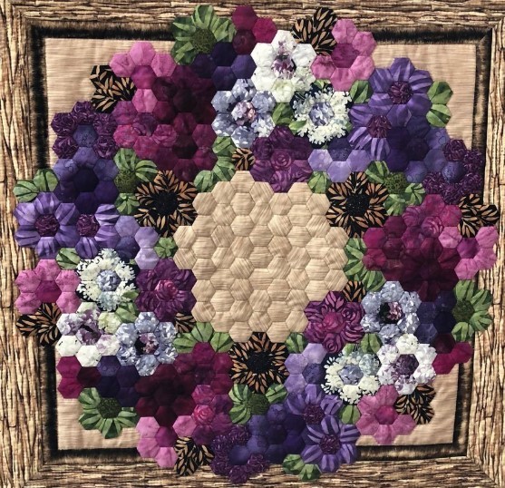 Hexagonal quilt featuring purple and pink fabric flowers arranged in wreath pattern with cream center and wood frame