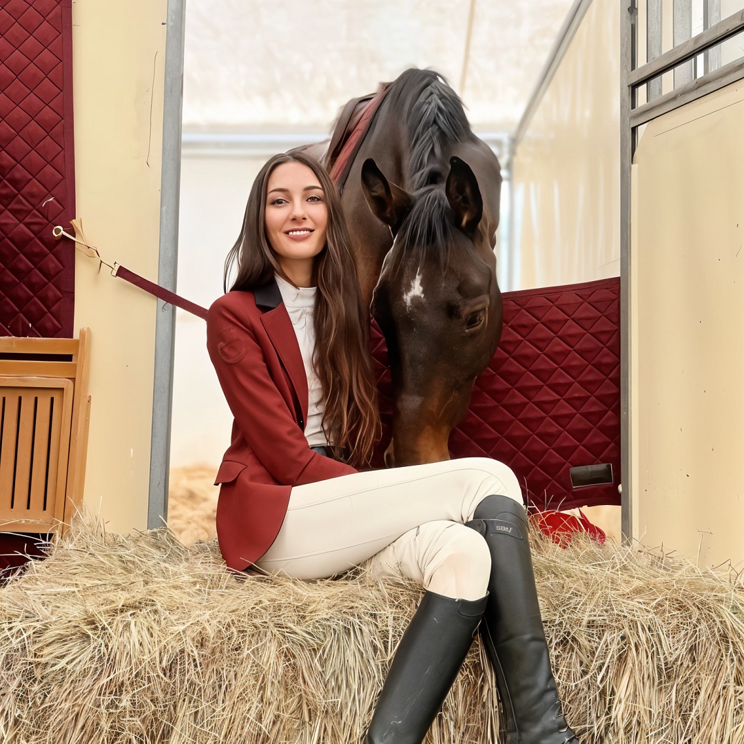 Woman in riding jacket and boots sitting next to brown horse in stable