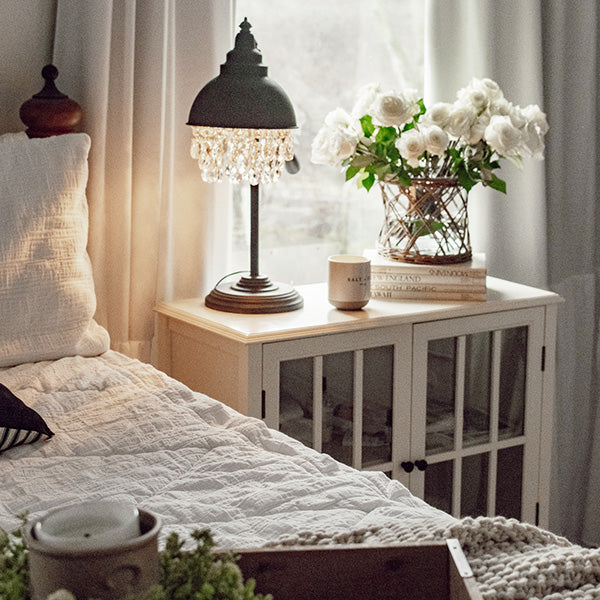 Cozy bedroom corner with white bedside table, lamp, flowers, bed, and sheer curtains