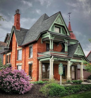 Red brick Victorian house with green ornamental trim, pointed gables, turet, and purple lilac bushes in front