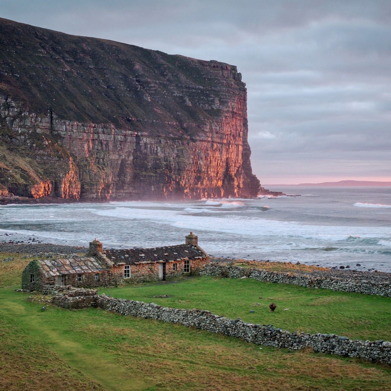 Stone cottage on grassy cliff overlooking beach with towering red rock cliffs at sunset in Orkney Scotland