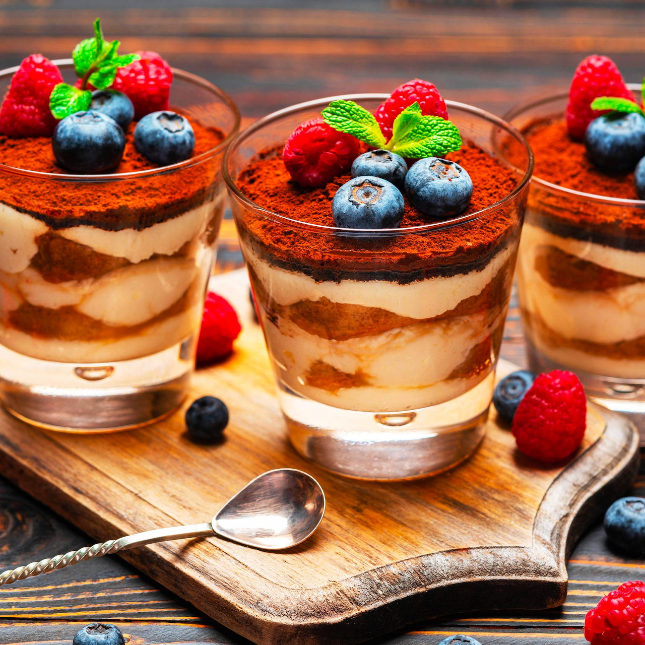 Three tiramisu dessert cups with berries and mint garnish on wooden board