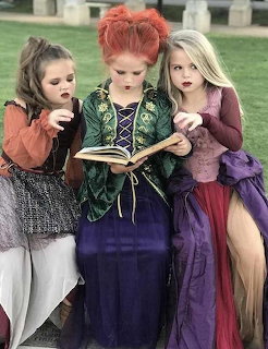 Three girls in Sanderson Sisters Halloween costumes holding a spell book on a grassy lawn
